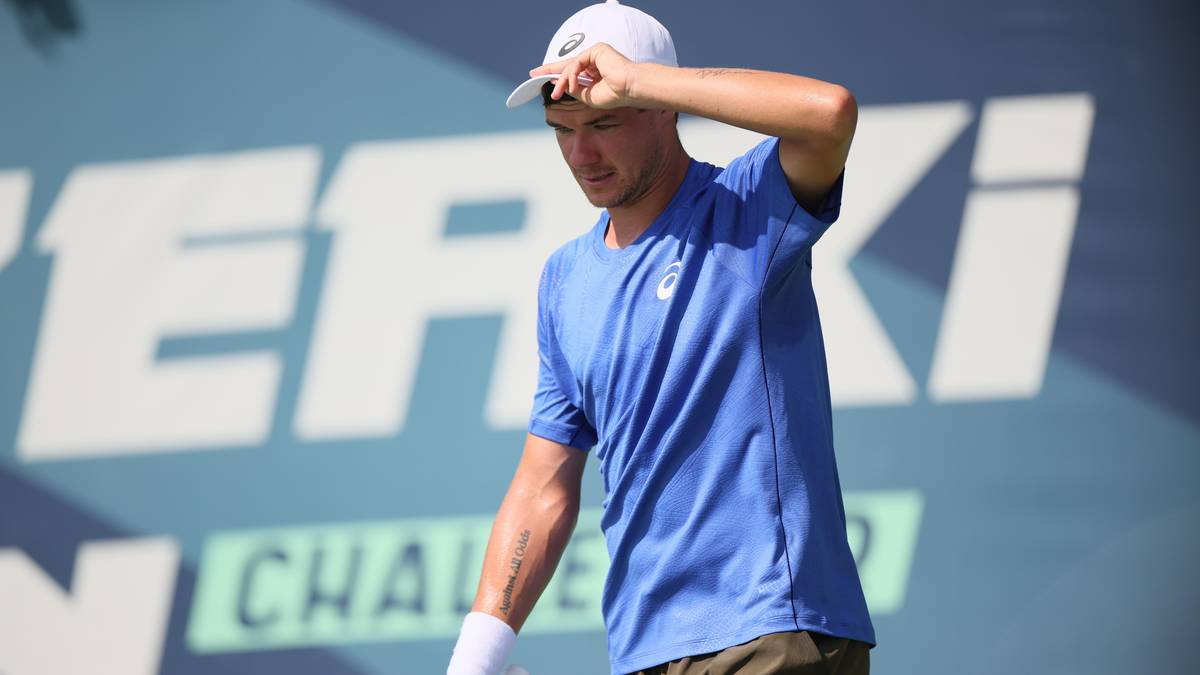 Młody mężczyzna w niebieskiej koszulce tenisowej i białej czapce zasłania oczy ręką, podczas meczu tenisowego.