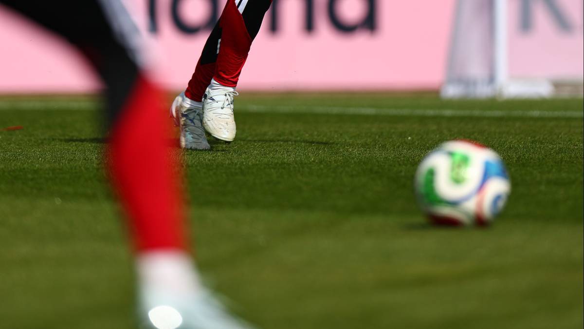 Zbliżenie na nogi piłkarza w czerwonych getrach i białe buty na zielonej murawie boiska piłkarskiego, w tle piłka nożna