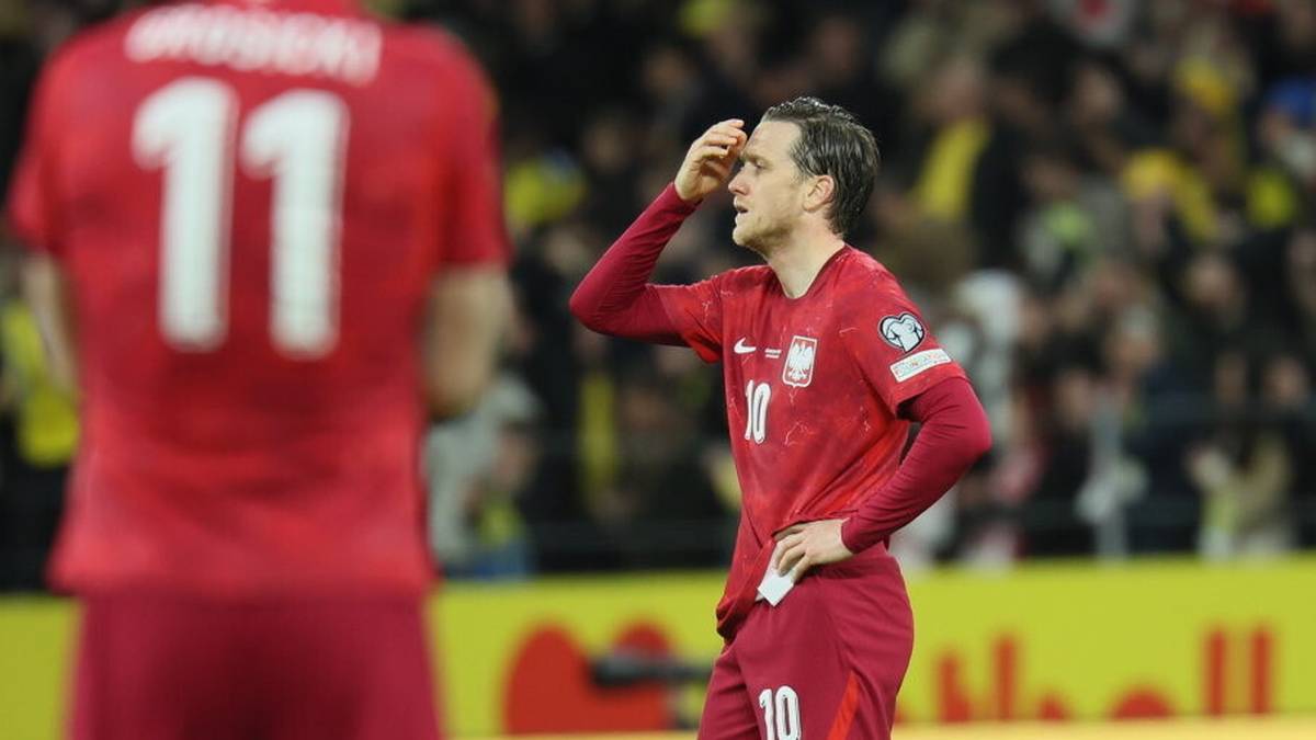 Piłkarz w czerwonej koszulce z numerem 10 na plecach, z dłonią przy czole, wyraża rozczarowanie na boisku piłkarskim.