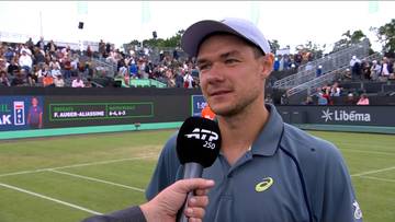 Kamil Majchrzak, polski tenisista, w czapce z daszkiem i koszulce sportowej, udziela wywiadu z mikrofonem ATP w ręce. Na jego twarzy widać uśmiech. W tle są trybuny z kibicami i kort tenisowy.