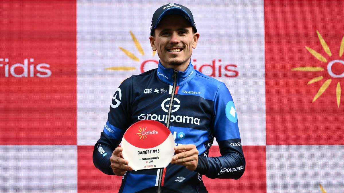 Kolarz w niebieskiej koszulce z logo Groupama-FDJ trzyma okrągłą nagrodę z emblematem słońca.