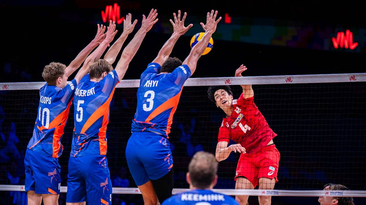 Gracze drużyny w niebieskich strojach próbują zablokować atak przeciwnika w czerwonym stroju podczas meczu siatkówki.