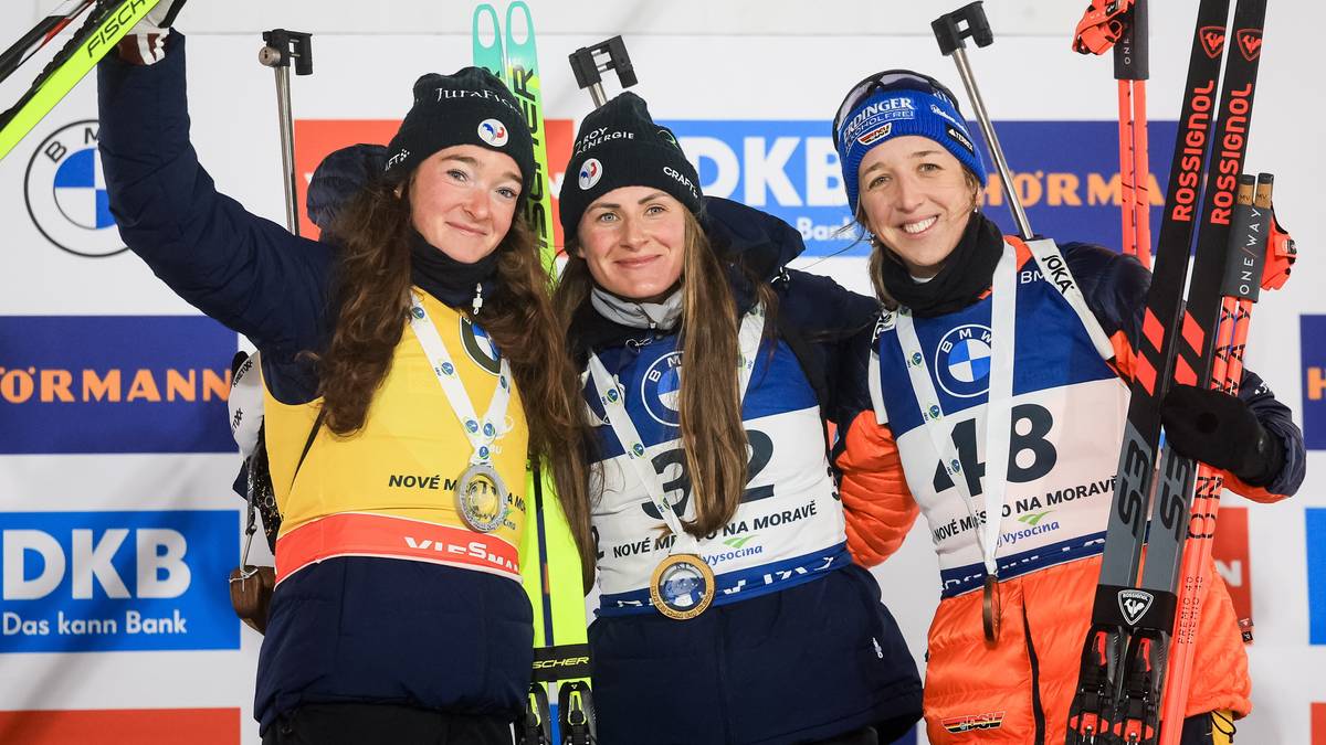 Trzy biathlonistki pozują do zdjęcia z medalami i nartami, świętując sukces na tle sponsor&oacute;w.