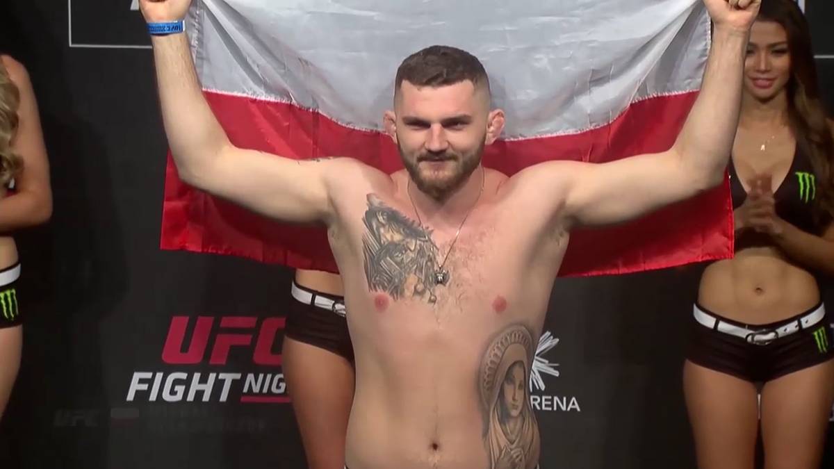 Mężczyzna z tatuażami na torsie trzyma polską flagę nad głową, stojąc na scenie UFC.