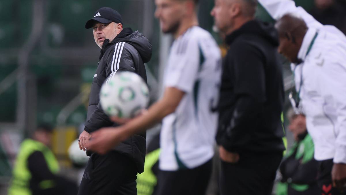 Trener Legii Warszawa w czapce z daszkiem i czarnej kurtce, patrzący w bok z poważną miną, trzymający piłkę.