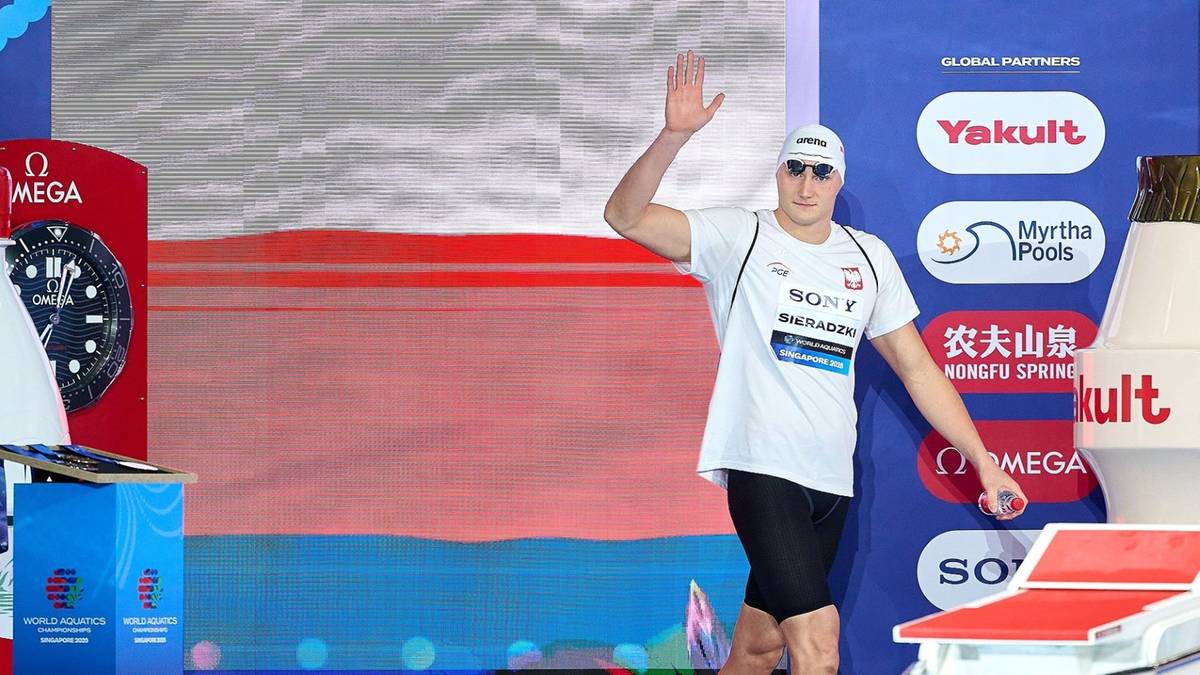 Pływak Kamil Sieradzki w stroju sportowym, z czapeczką i okularami, podnosi prawą rękę do góry w geście pozdrowienia. Na piersiach ma przypięty numer startowy i widnieją logotypy sponsorów. W tle znajdują się ekrany z wyświetlanymi informacjami, strefa dla widzów i fragmenty toru pływackiego.