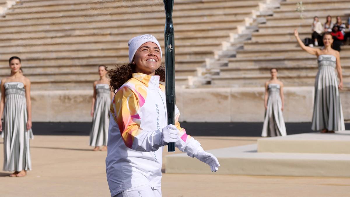 Uśmiechnięta młoda kobieta w białym stroju z kolorowymi rękawami i czapką trzyma pałkę w rękach, stojąc na tle trybun stadionu.