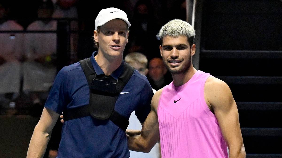 Dw&oacute;ch mężczyzn, tenisist&oacute;w, stoją obok siebie, jeden w niebieskiej koszulce i czapce, drugi w r&oacute;żowej koszulce, obaj uśmiechają się.