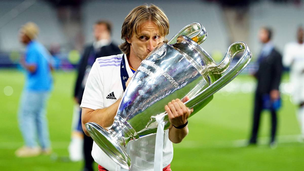 Piłkarz Luka Modrić trzyma w ramionach duże, srebrne trofeum, całując je. Jest w białej koszulce z logo Realu Madryt. W tle widać innych ludzi i zieloną murawę stadionu.