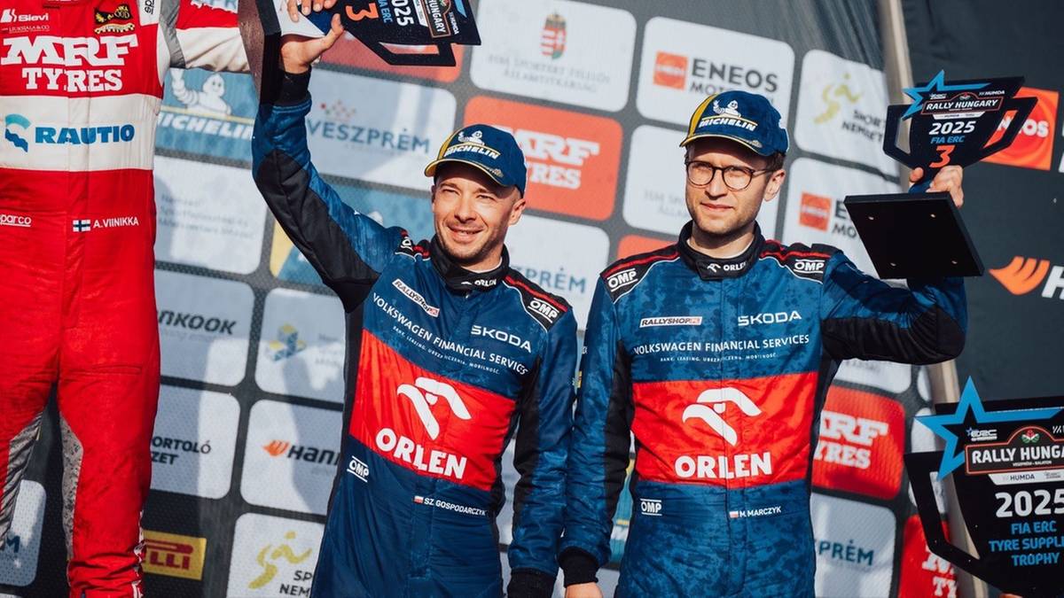 Dwóch mężczyzn w kombinezonach rajdowych z napisem ORLEN na piersiach, trzymających puchary, z sukcesem pozujących do zdjęcia na tle sponsorów.