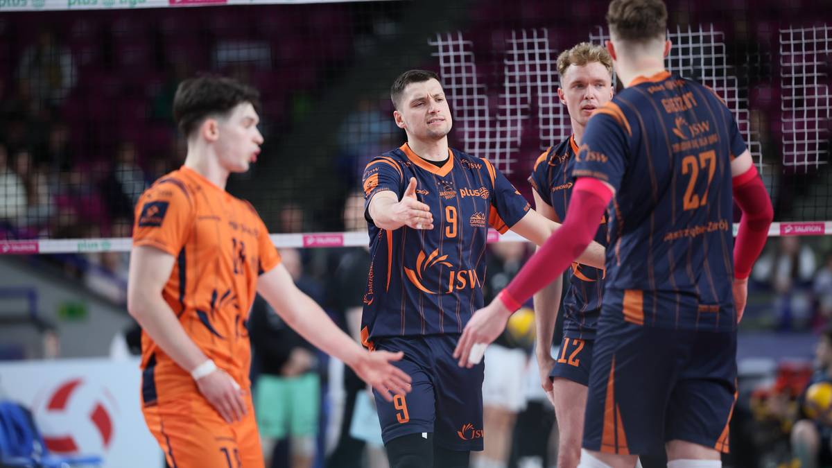 Czterech siatkarzy na boisku podczas meczu siatk&oacute;wki. Dw&oacute;ch zawodnik&oacute;w w pomarańczowych strojach nawiązuje kontakt fizyczny, pozostali dwaj w granatowych strojach stoją obok. Siatka do siatk&oacute;wki i fragment trybun są widoczne w tle.