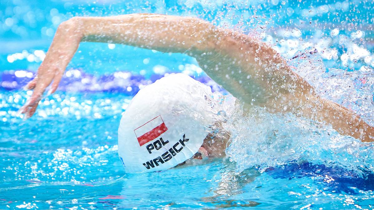 Pływak w czepku z polską flagą wykonuje ruch ręką w wodzie.