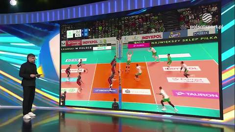 Duży ekran prezentujący mecz siatk&oacute;wki z graczami w akcji, boiskiem i trybunami pełnymi kibic&oacute;w. Obok ekranu stoi mężczyzna w czarnym garniturze.