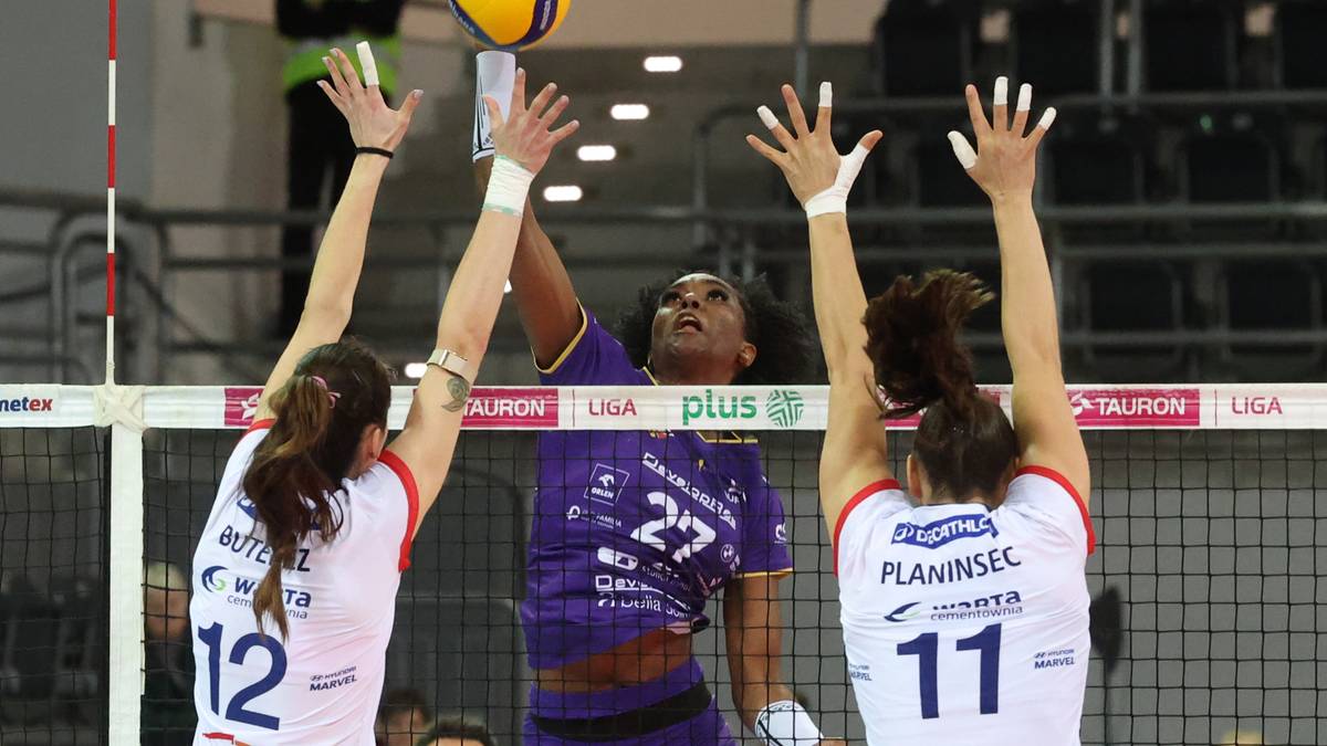 Trzy siatkarki w akcji przy siatce. Dwie zawodniczki w białych strojach pr&oacute;bują zablokować atak zawodniczki w fioletowym stroju, kt&oacute;ra uderza w piłkę.