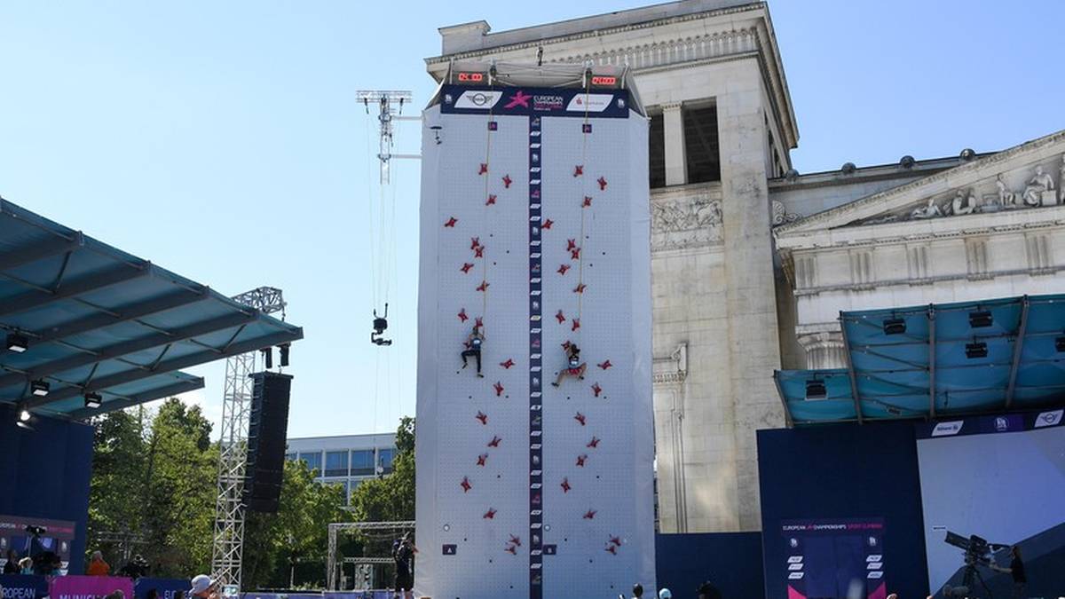 Wysoka, biała ścianka wspinaczkowa z różowymi chwytami, na której znajdują się dwie wspinające się osoby.