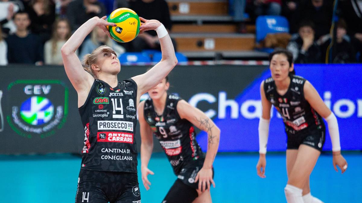 Siatkarka w czarnym stroju z numerem 14 na plecach, z piłką do siatkówki w rękach, przygotowuje się do zagrywki. W tle widać inne zawodniczki oraz trybuny z widzami.