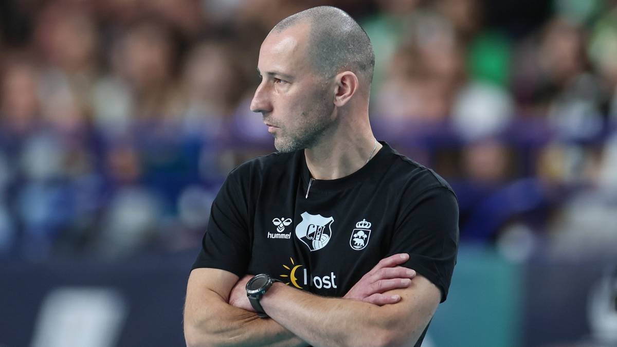 Mężczyzna w czarnej koszulce sportowej z logo klubu, stoi ze skrzyżowanymi ramionami i patrzy w bok.