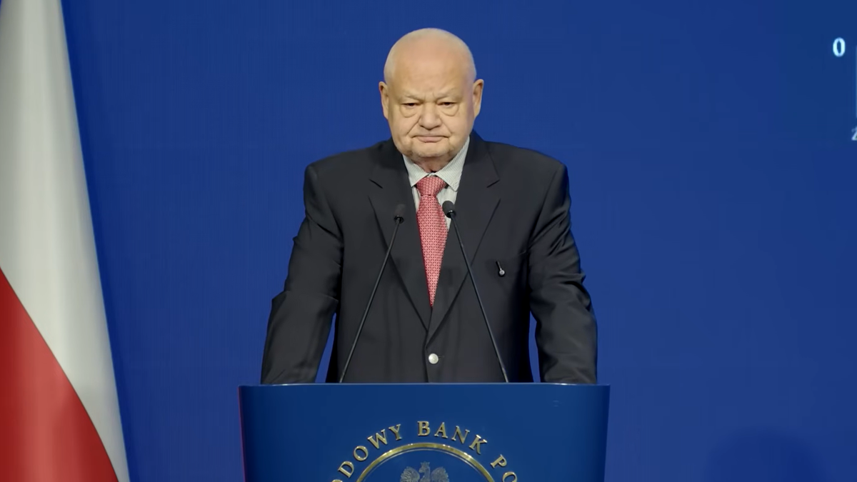 Starszy mężczyzna w garniturze stoi za m&oacute;wnicą Narodowego Banku Polskiego.