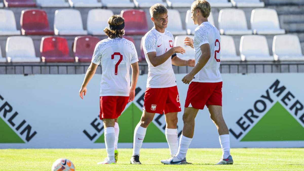Trzech młodych piłkarzy w biało-czerwonych strojach, stojących na boisku, jeden z nich rozmawia z drugim.