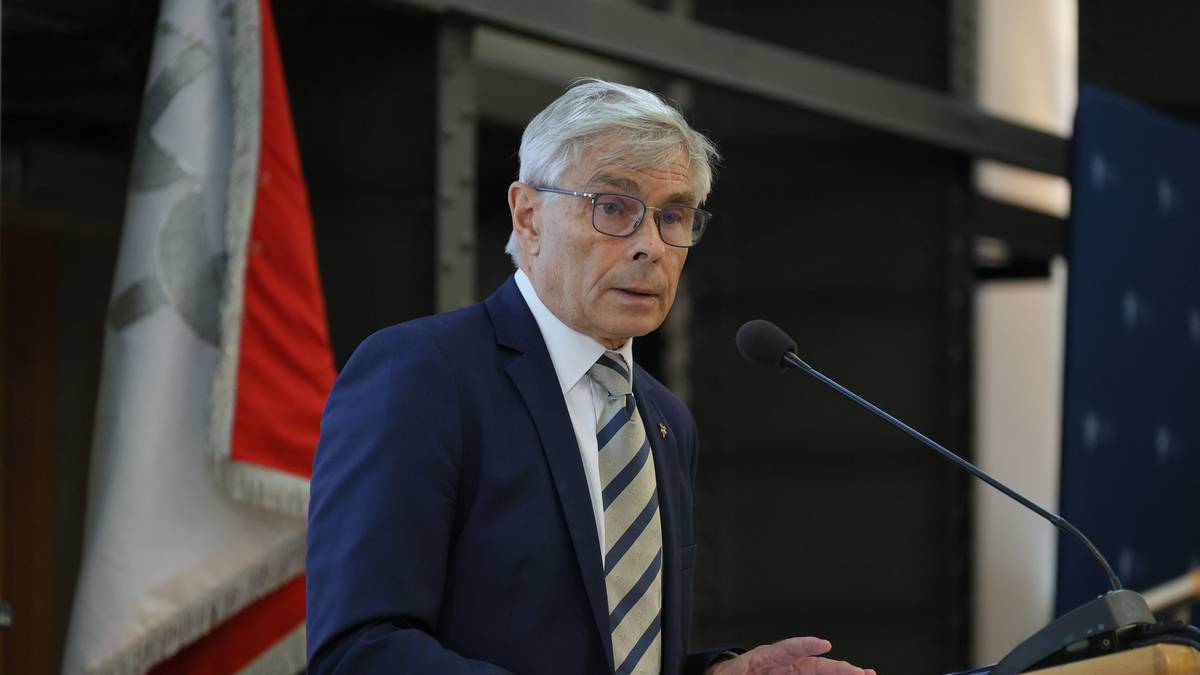 Starszy mężczyzna w okularach i garniturze przemawia za mównicą, obok widoczna flaga.