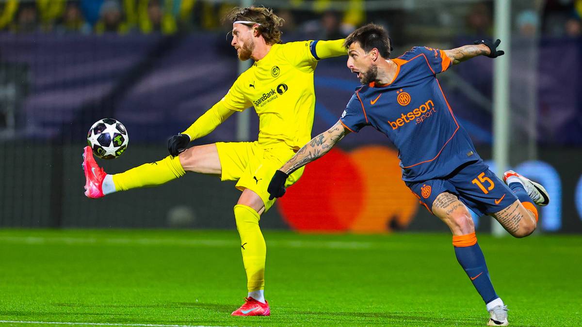 Dw&oacute;ch piłkarzy na boisku piłkarskim, jeden w ż&oacute;łtym stroju kopie piłkę, drugi w granatowym pr&oacute;buje mu przeszkodzić.