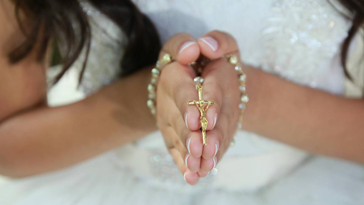Dłonie dziecka trzymające złoty r&oacute;żaniec ze złotym krzyżykiem, złożone do modlitwy.