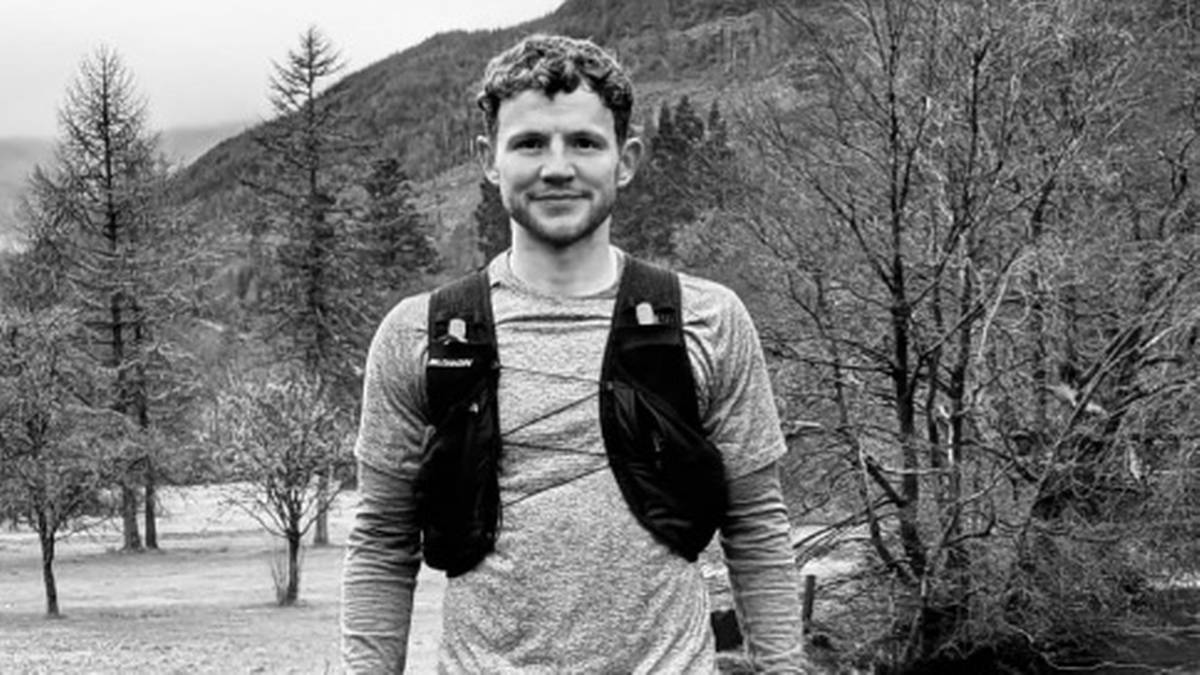 Uśmiechnięty mężczyzna w koszulce z długim rękawem i kamizelce biegowej stoi na zewnątrz, z drzewami i wzg&oacute;rzami w tle.