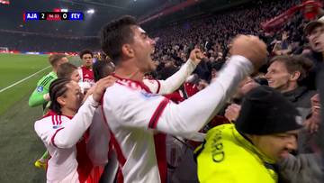 Piłkarz Ajaxu Amsterdam w białej koszulce z czerwonymi elementami świętuje z innymi zawodnikami i kibicami.