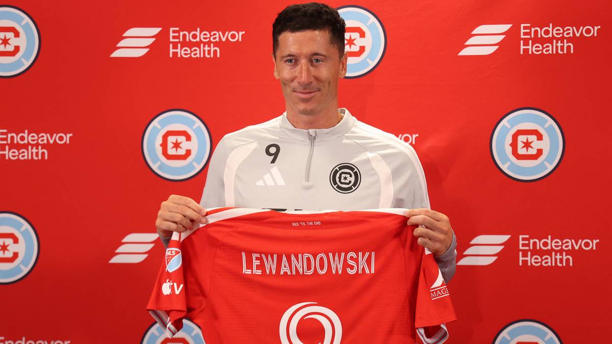 Robert Lewandowski trzyma czerwono-białą koszulkę piłkarską z napisem "LEWANDOWSKI" na plecach, stojąc przed ścianką z logo "Endeavor Health" i symbolem "C" w okręgu.