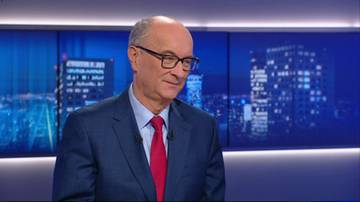 Starszy mężczyzna w okularach i garniturze z czerwonym krawatem siedzi przed ekranami z nocnym widokiem miasta.