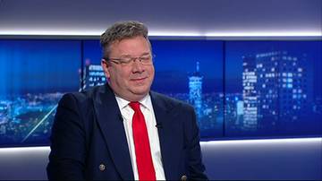 Mężczyzna w garniturze i czerwonym krawacie siedzi przed ekranem przedstawiającym nocną panoramę miasta.