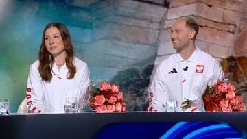 Kobieta i mężczyzna siedzą przy stole, uśmiechają się. Przed nimi stoją kwiaty i szklanki.