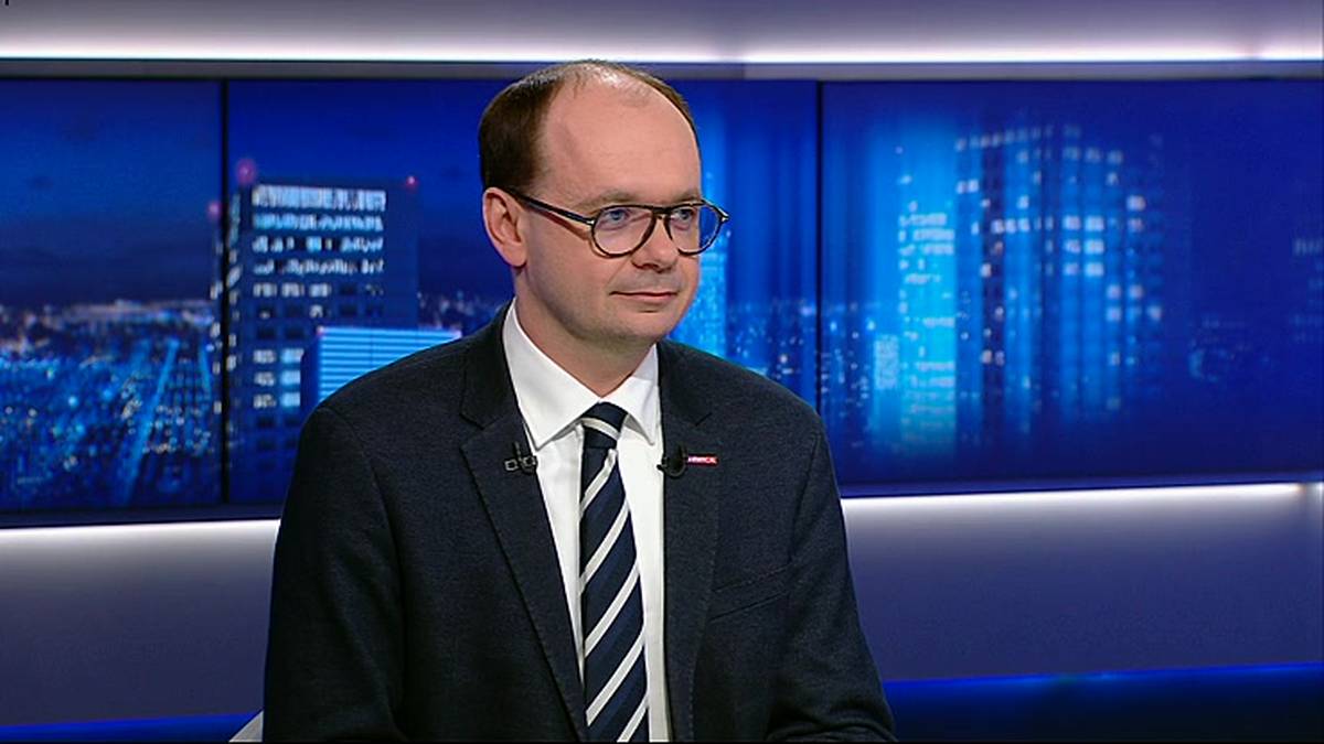 Mężczyzna w okularach, białej koszuli i krawacie w paski, ubrany w ciemny garnitur, siedzi przed ekranem z widokiem nocnego miasta.