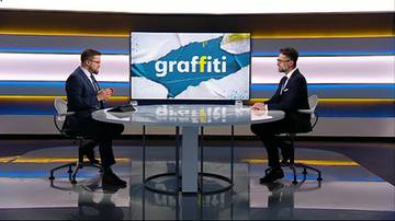 Na ekranie widnieje napis "graffiti" na niebieskim tle z białym efektem pęknięcia. Dw&oacute;ch mężczyzn w garniturach siedzi naprzeciwko siebie przy okrągłym stole w studiu telewizyjnym.
