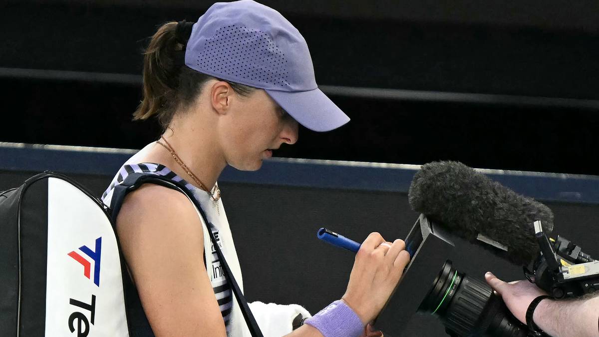 Iga Świątek w czapce z daszkiem siedzi i podpisuje coś na kartce, obok niej mikrofon kamery.