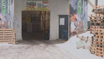 Wejście do magazynu z paletami pelletu opałowego, stosy palet na zewnątrz, banery z napisem PELLET, śnieg na ziemi.