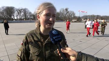 Kobieta w mundurze wojskowym, Katarzyna Michałowska-Dąbrowska, udziela wywiadu mikrofonowi z logo Polsat Sport. W tle widać plac, innych żołnierzy w strojach sportowych oraz biało-czerwone flagi Polski na masztach.