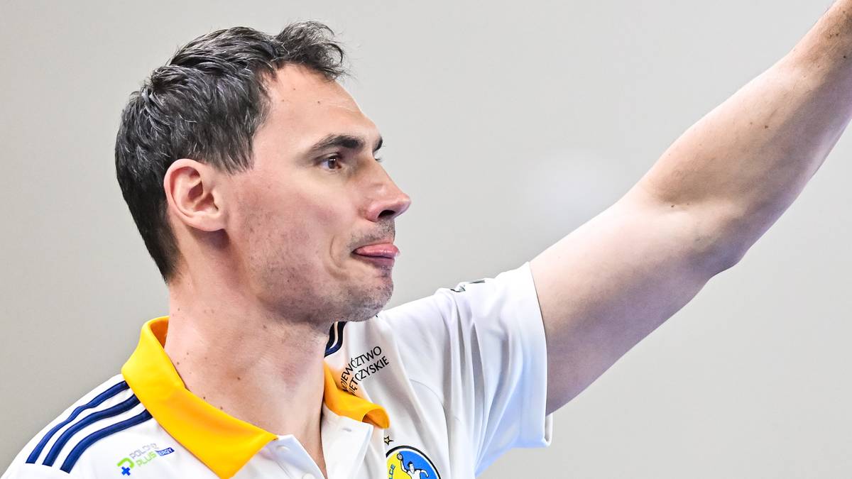 Mężczyzna w białej koszulce sportowej z ż&oacute;łtym kołnierzykiem unosi prawą rękę, wskazując palcami do g&oacute;ry.