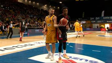 Dw&oacute;ch koszykarzy stoi na boisku, jeden w ż&oacute;łtym, drugi w czarnym stroju, z piłką między nimi.