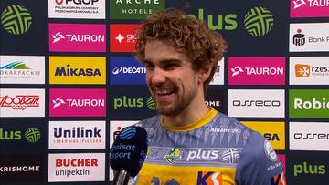 Młody mężczyzna z kręconymi blond włosami, uśmiechnięty, w bluzie sportowej, stoi przed ścianą z logo sponsor&oacute;w.