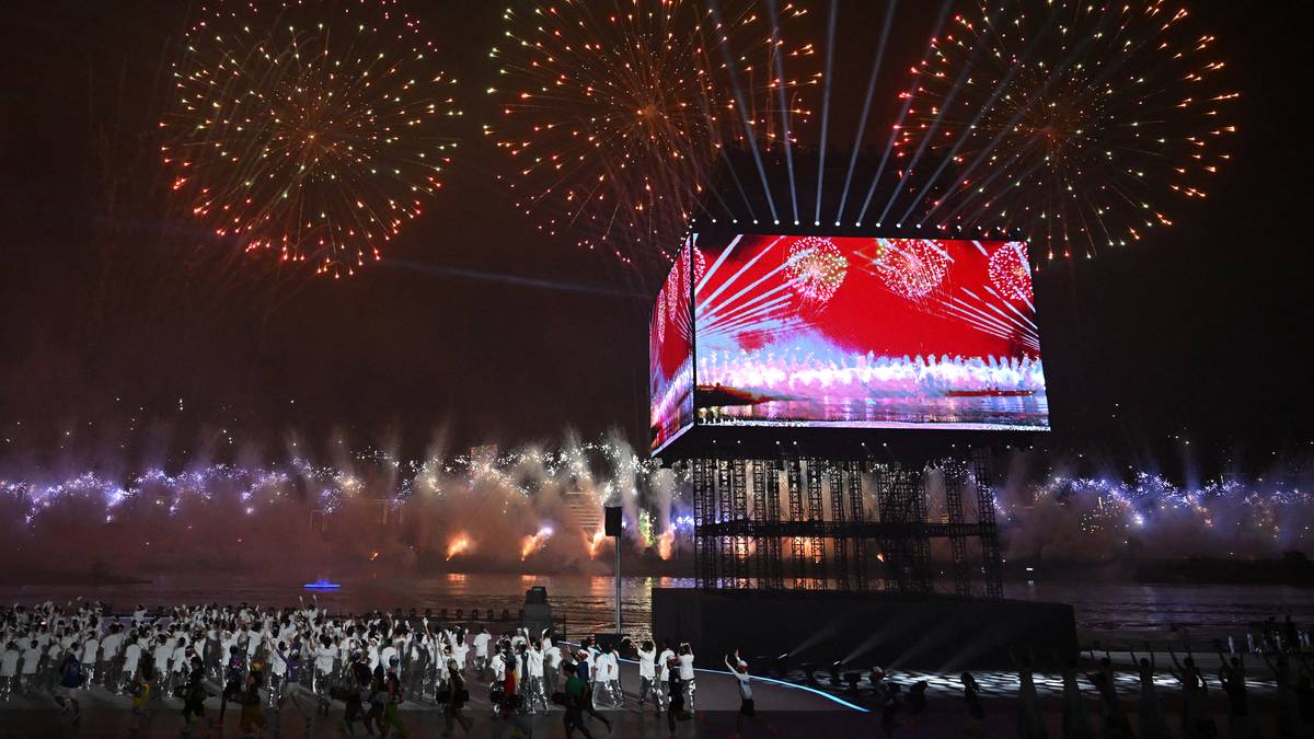 Wielki ekran wyświetlający jasne, kolorowe wzory na tle nocnego nieba, nad którym odbywa się pokaz sztucznych ogni. Na dole widoczny jest duży tłum ludzi zgromadzony na otwartej przestrzeni.