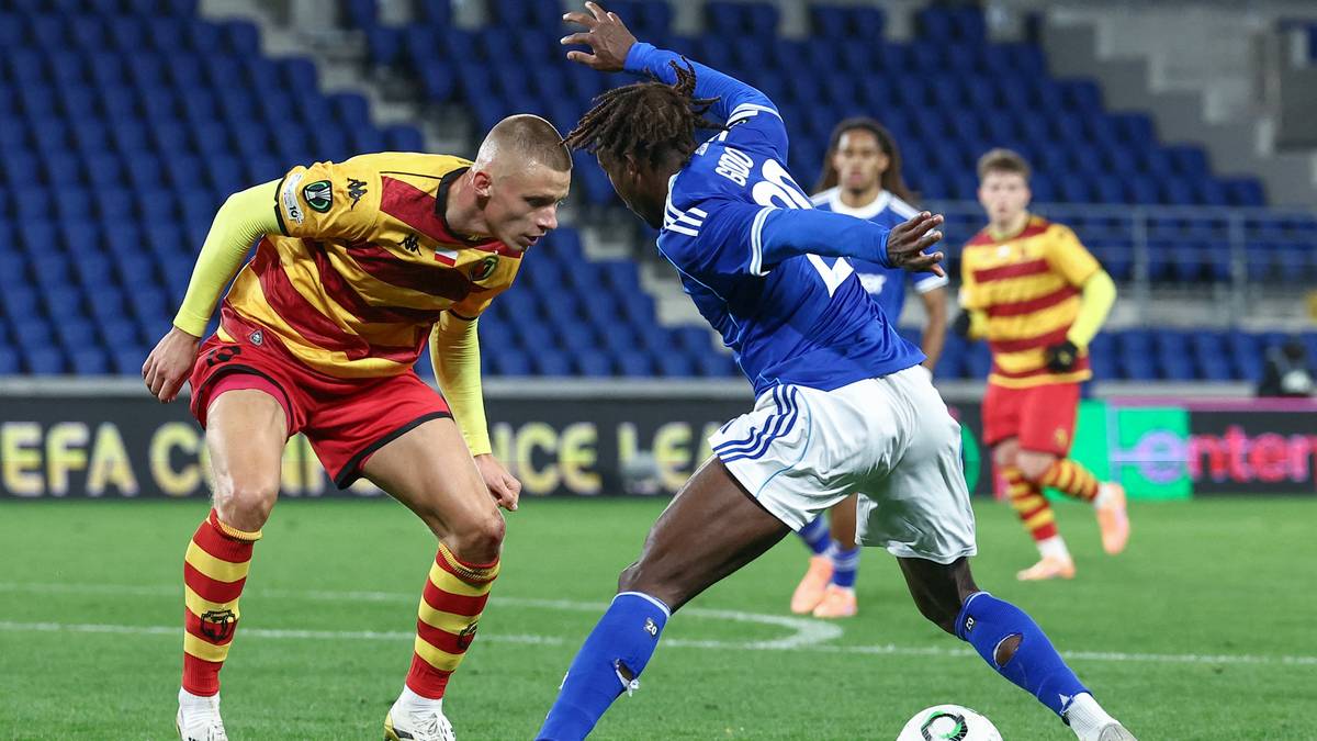 Dwóch piłkarzy podczas meczu, jeden w żółto-czerwonym stroju, drugi w niebieskim, walczą o piłkę na boisku.