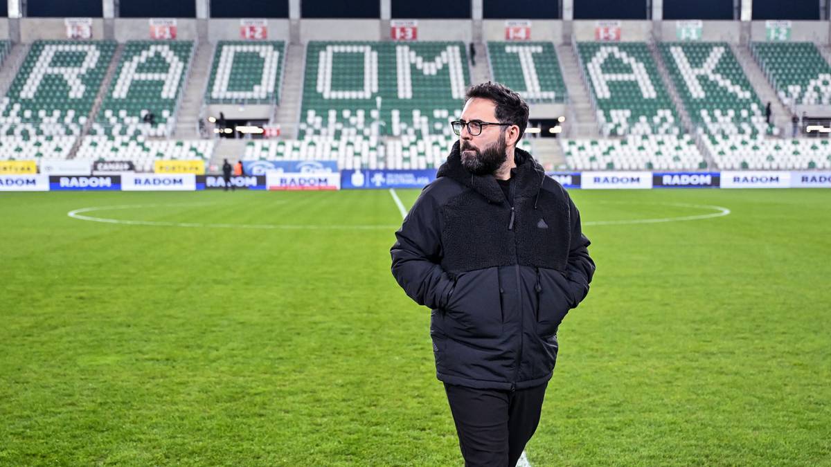 Mężczyzna w ciemnej kurtce i okularach stoi na boisku piłkarskim przed trybunami stadionu z napisem RADOMIAK.