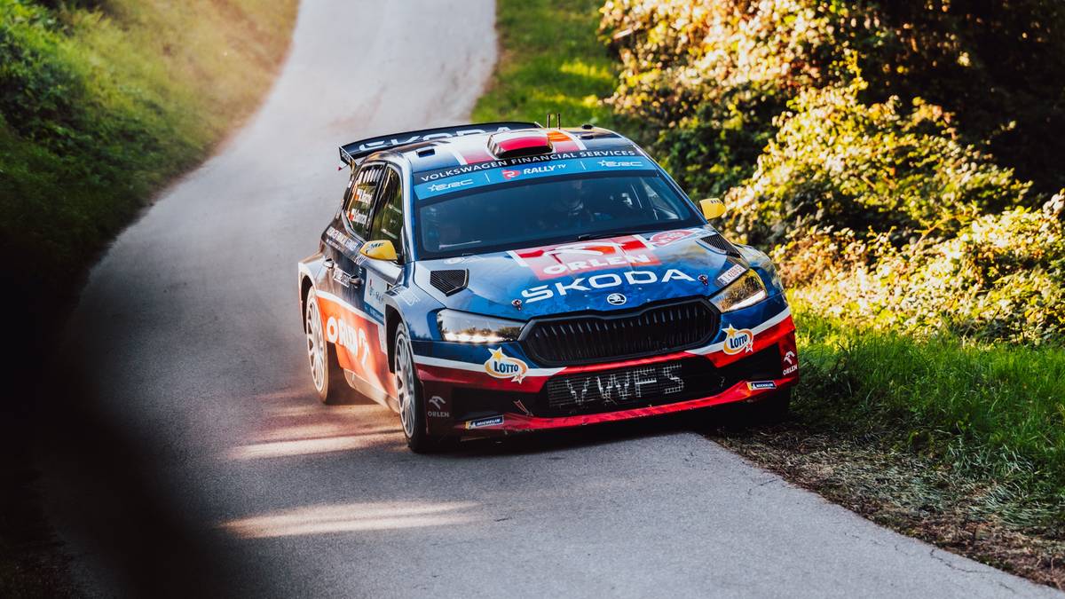 Samochód rajdowy Skoda Fabia w barwach niebieskich, czerwonych i białych jedzie po wąskiej, krętej drodze otoczonej przez zielony las. Na masce widnieje napis "SKODA". Kierownica widoczna jest przez szybę.