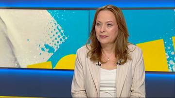 Kobieta w jasnej marynarce siedzi przed ekranem z abstrakcyjnym tłem.
