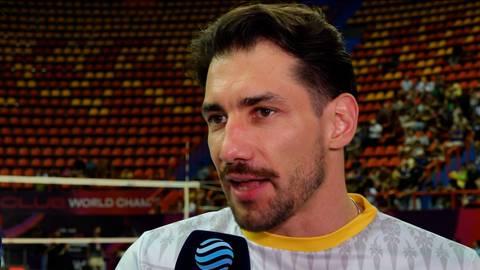 Portret mężczyzny w stroju sportowym, prawdopodobnie siatkarza, udzielającego wywiadu. W tle widoczne są trybuny stadionu.