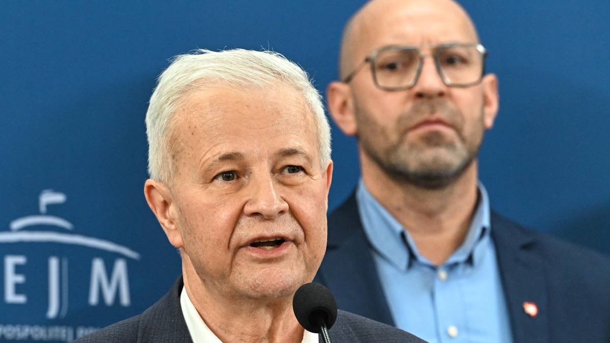 Starszy mężczyzna o siwych włosach przemawia, za nim stoi mężczyzna w okularach.