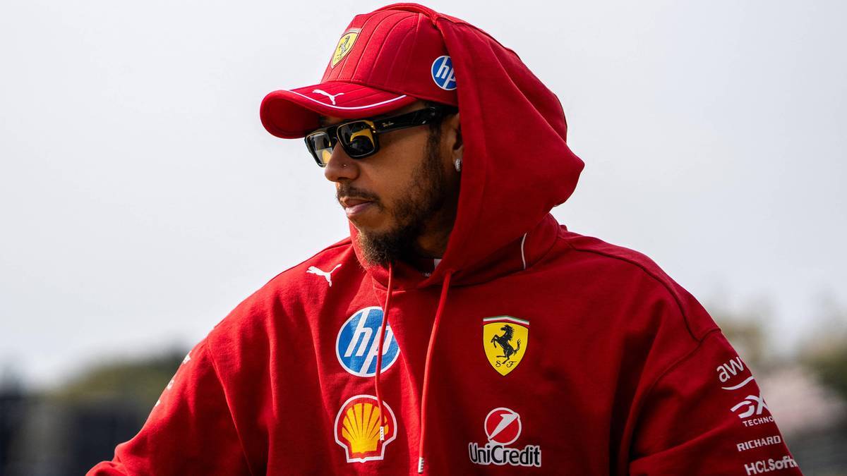 Lewis Hamilton w czerwonej bluzie z kapturem i czapką Ferrari, noszący okulary przeciwsłoneczne.