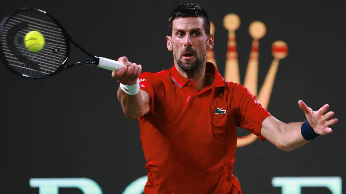 Novak Djoković uderza piłkę rakietą tenisową podczas meczu. Ma na sobie czerwoną koszulkę polo z logo Lacoste i białą opaskę na nadgarstku. W tle widoczne jest logo Rolex.
