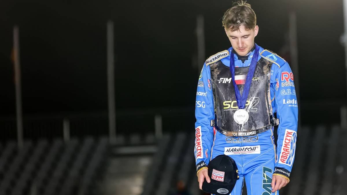 Młody mężczyzna w kombinezonie żużlowym, z medalem na szyi i czapką w dłoni, patrzy w dół ze smutnym wyrazem twarzy.